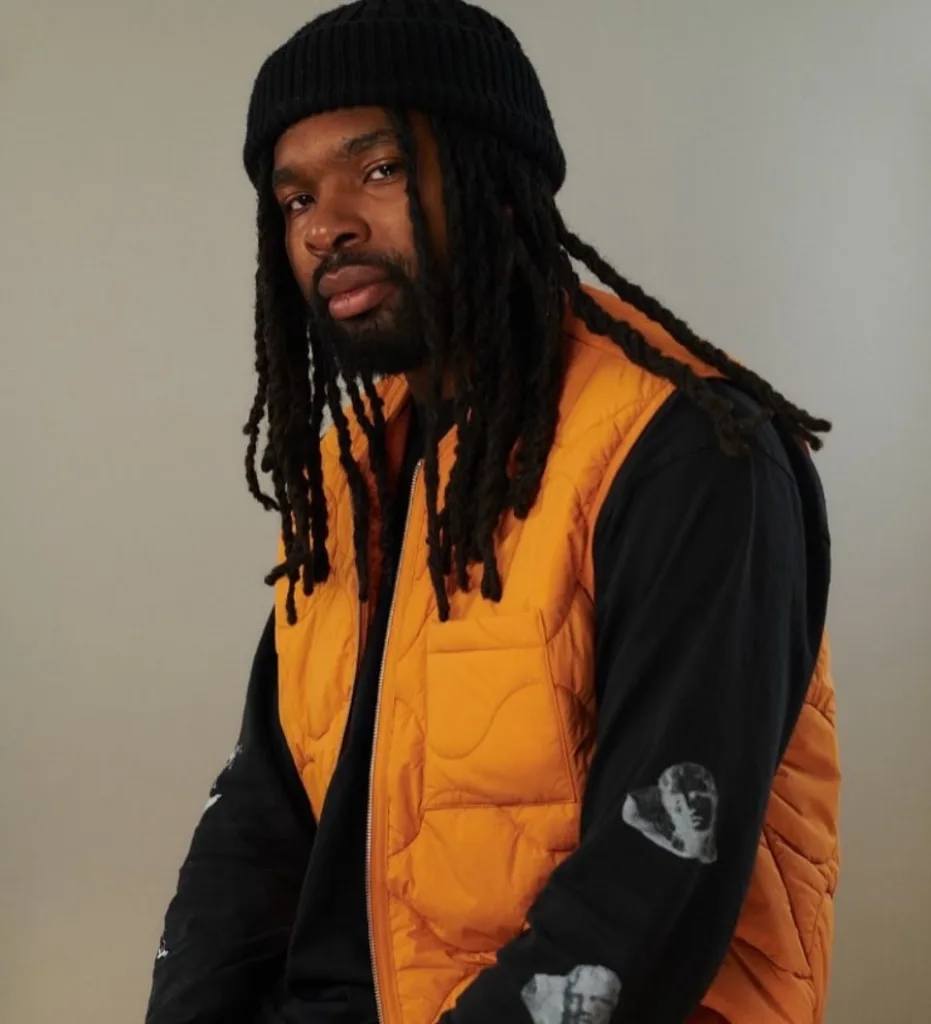 ItsNate with long locks and a beard wearing a black beanie and orange padded gilet over black clothing